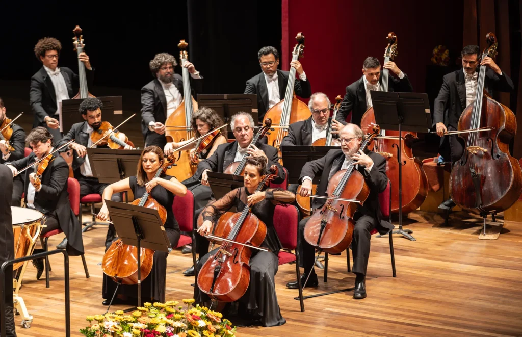 Imagem da Orquestra Sinfônica Brasileira tocando no Teatro Municipal, onde aparecem 13 músicos com diferentes.